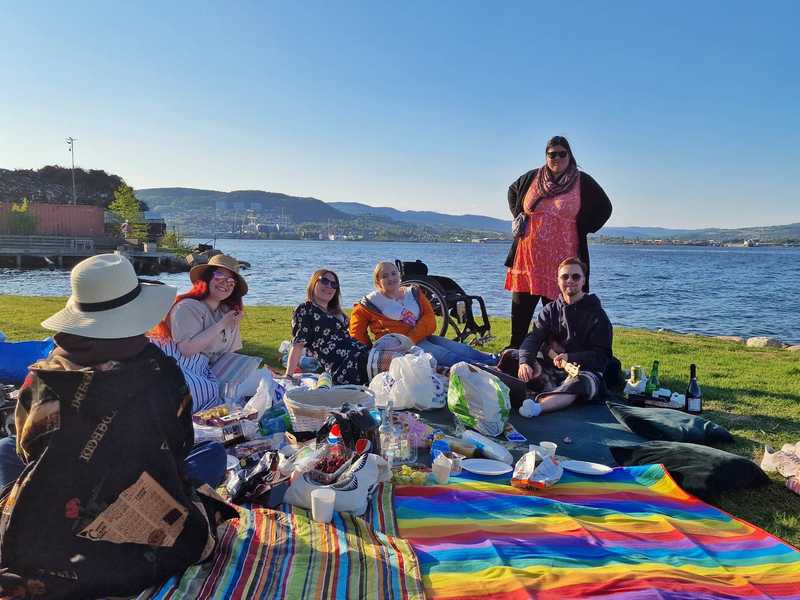 6 personer sitter på tepper på gresset ved drammenselva. Det er en piknikk, med mange fargefulle tepper, mat, drikke og en høytaler. Det er sol og godt sommervær, og nesten alle i bildet har på solbriller og en solhatt. De smiler og ser mot kameraet som tar dette bildet. Folk sitter nærme hverandre, noen med et glass i hånden. En rullestol står rett bak en person