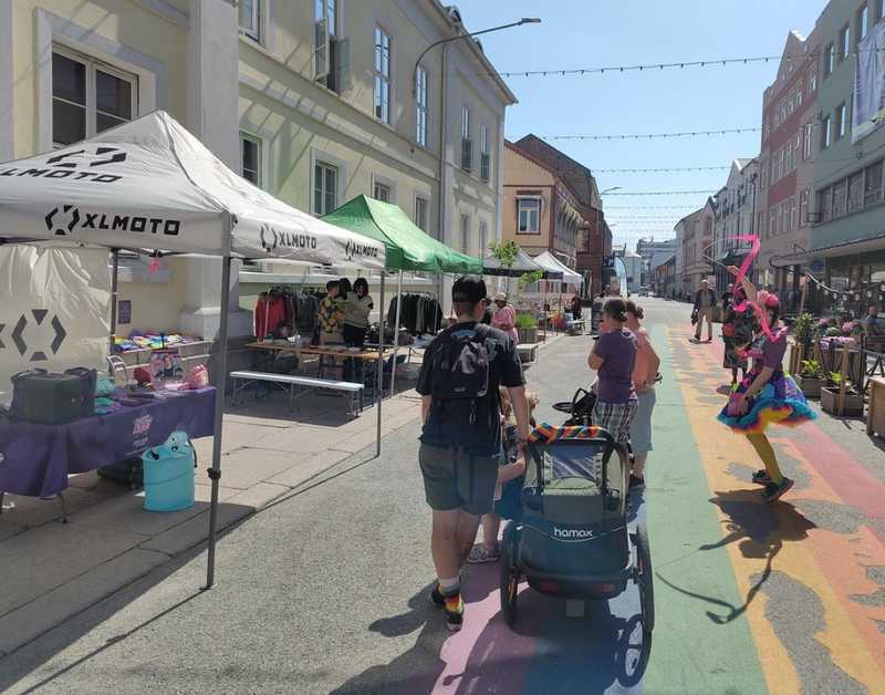 Bilde av et marked med flere bemannede boder og personer som går gjennom gaten markedet er på. Selve gaten er malt med en lang regnbue. Det er en klovn som lager show. Det er mange prideflagg i bakgrunnen.