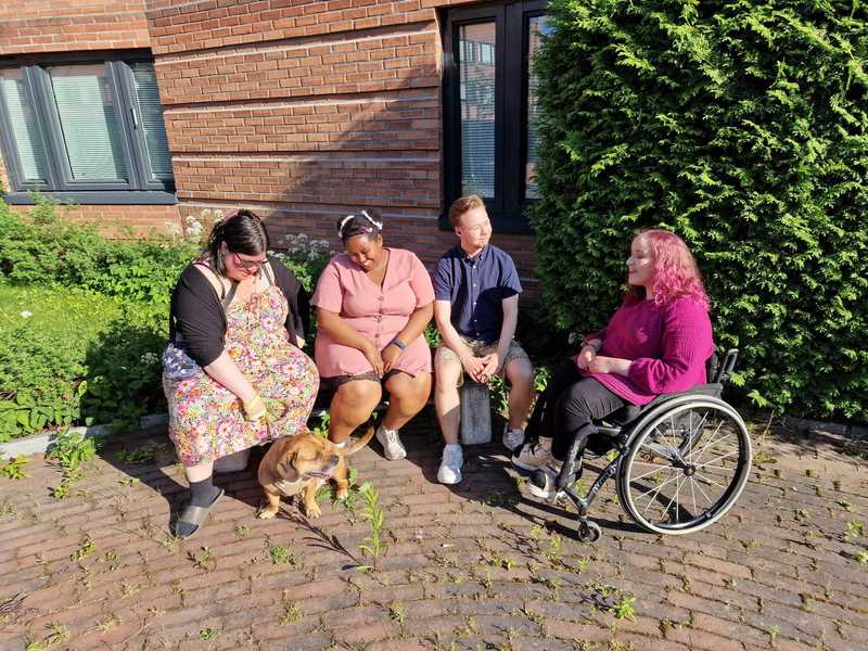 4 people are sitting on a bench out in the sun. A dog is also with them. Everyone is smiling and looking at each other, generally hanging out. One is in a wheelchair.